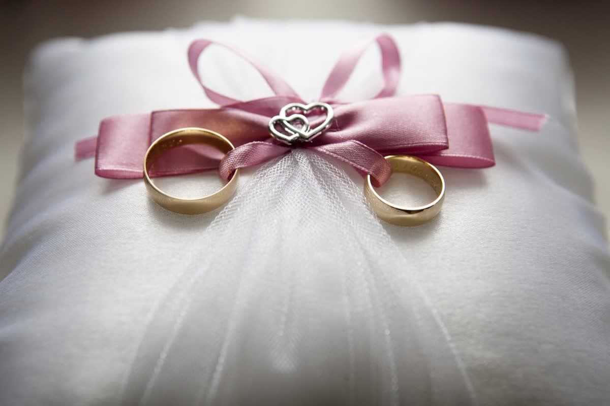 Two gold wedding rings on a white cushion with a pink ribbon